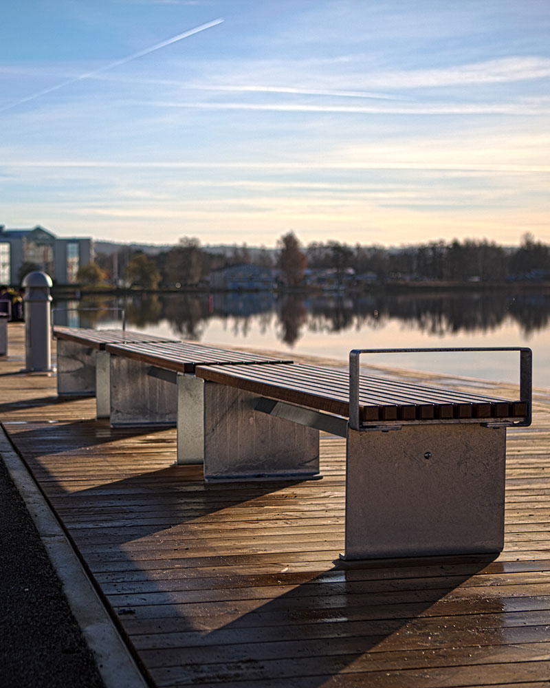 Bancs modulaires reliés entre eux le long d'une promenade au bord d'un lac en Suède, avec un soleil couchant en arrière-plan.